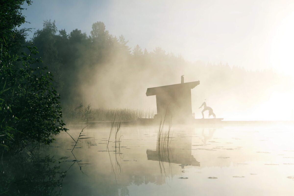 Person practicing yoga on a lakeside dock beside a small sauna hut in misty, calm water surrounded by forest.