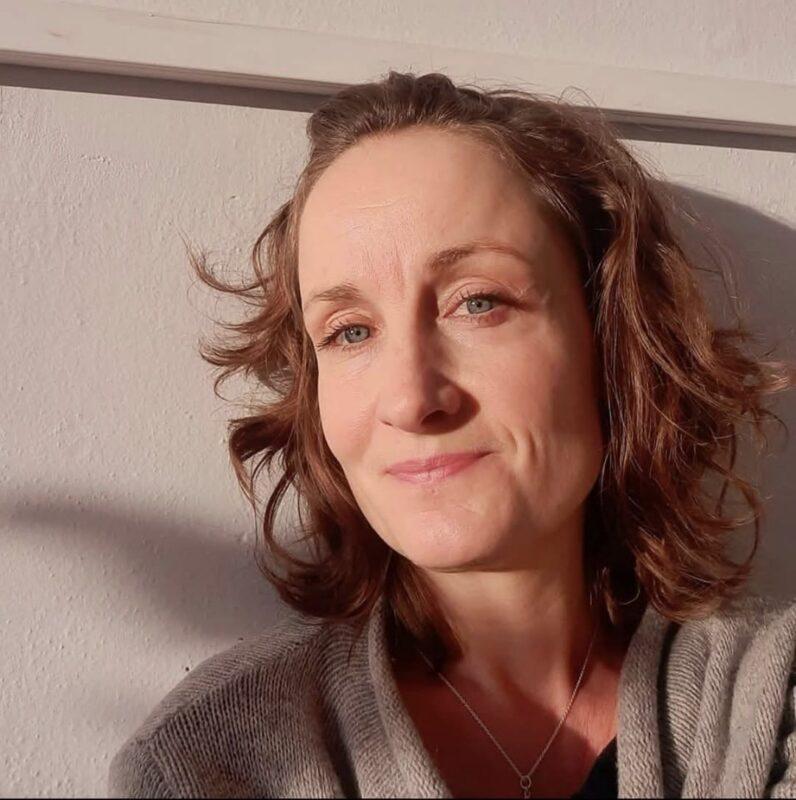 Portrait of a woman with wavy brown hair, softly lit by warm sunlight, smiling gently against a light-colored wall.