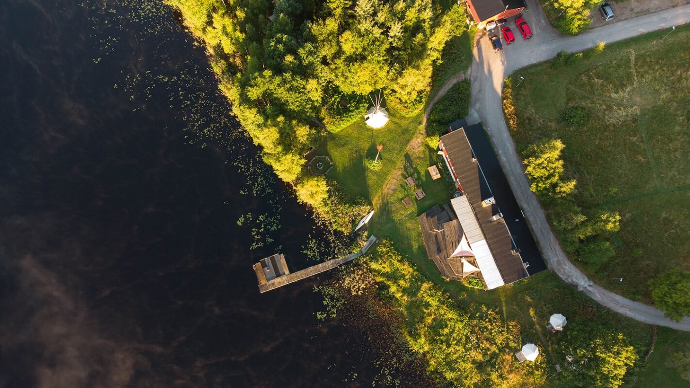 Aerial view of a lakeside retreat center with a dock, sauna, forest, and buildings surrounded by greenery.