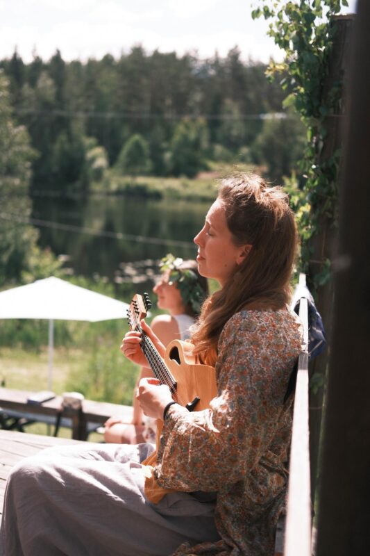 A woman sitting outdoors on a sunny wooden deck, peacefully playing a small string instrument with a lake and forest in the background. A calm and intimate retreat atmosphere.