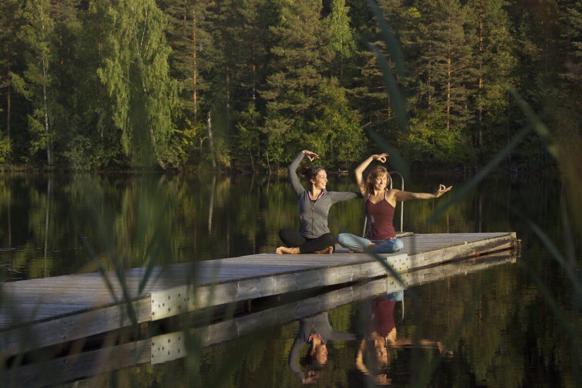 Women practicing yoga and meditation on a lakeside dock surrounded by forest at Shambala Gatherings