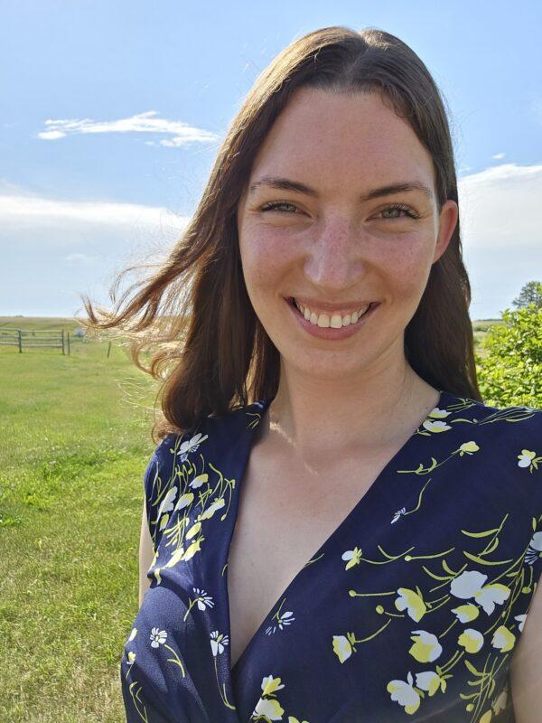 A smiling woman with long brown hair stands outdoors in a sunny grassy field, wearing a navy blue floral dress. The photo is backlit by bright sunlight, with a wide open landscape and dramatic blue sky in the background.