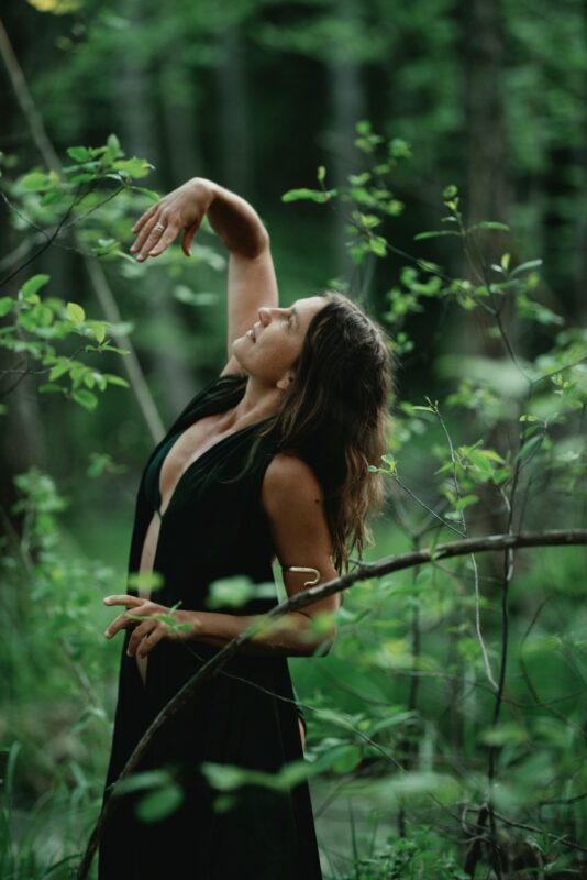 A woman moving gracefully among the trees in a green forest, with one arm lifted and a peaceful smile on her face.