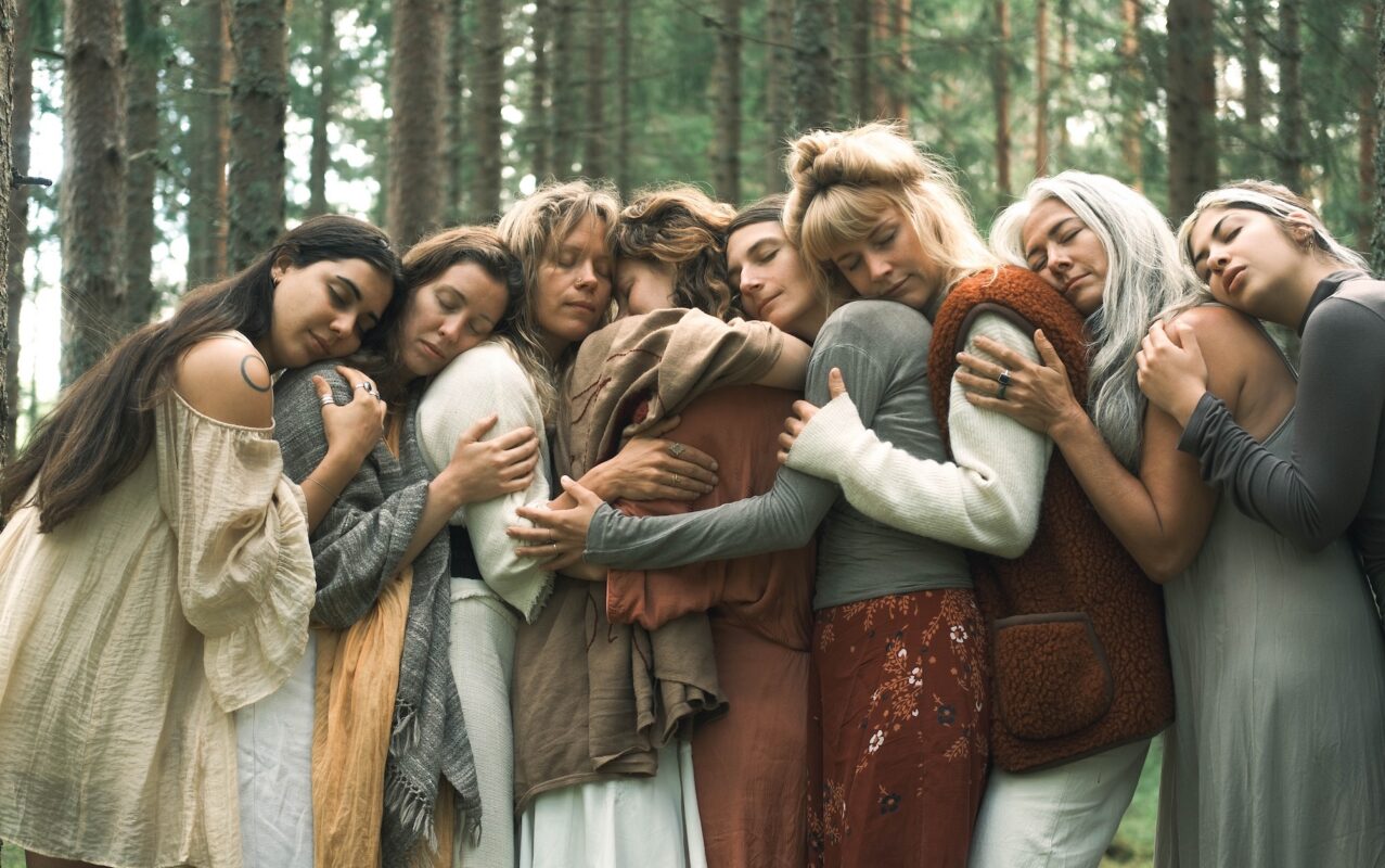 A group of seven women standing closely together in a forest, embracing one another with their eyes closed. The women are dressed in cozy, natural-toned clothing, creating a warm and intimate sense of connection and sisterhood.
