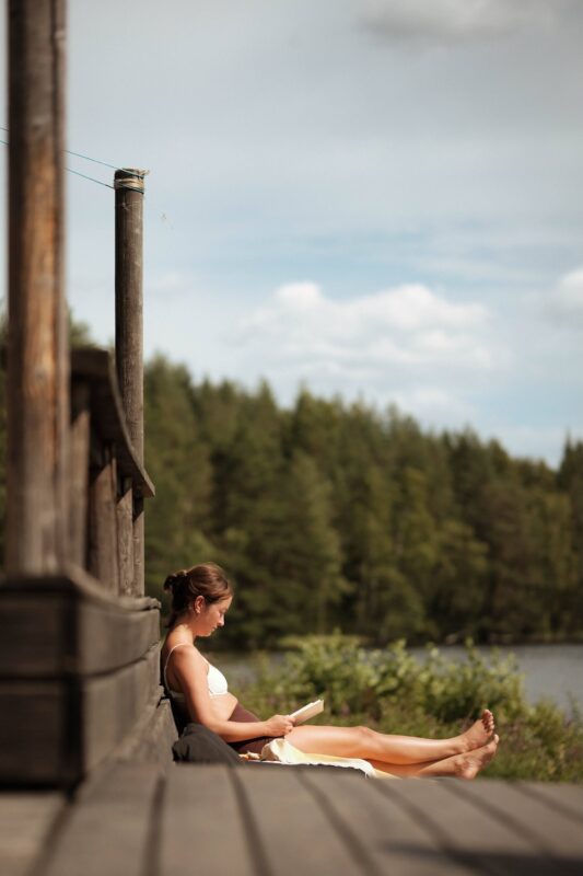 A person sitting on a wooden deck by the lake, reading a book with a forest in the background.